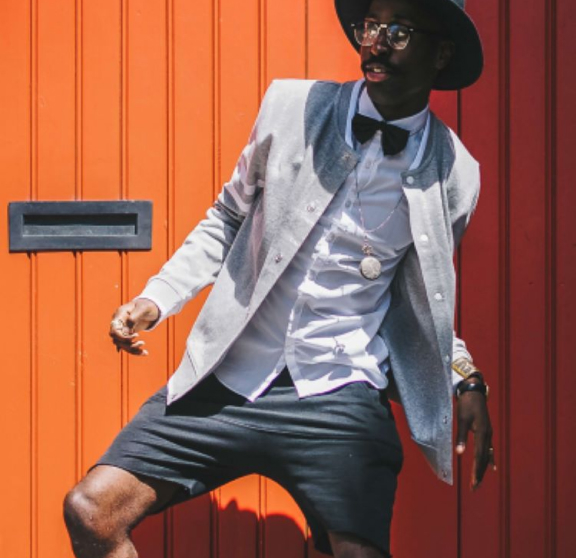 Modelo masculino vestindo chapéu preto, blazer cinza, camisa social branca, gravata borboleta na cor preta, óculos com lente transparente e armação na cor preta, além de um cordão prateado, em um fundo laranja.